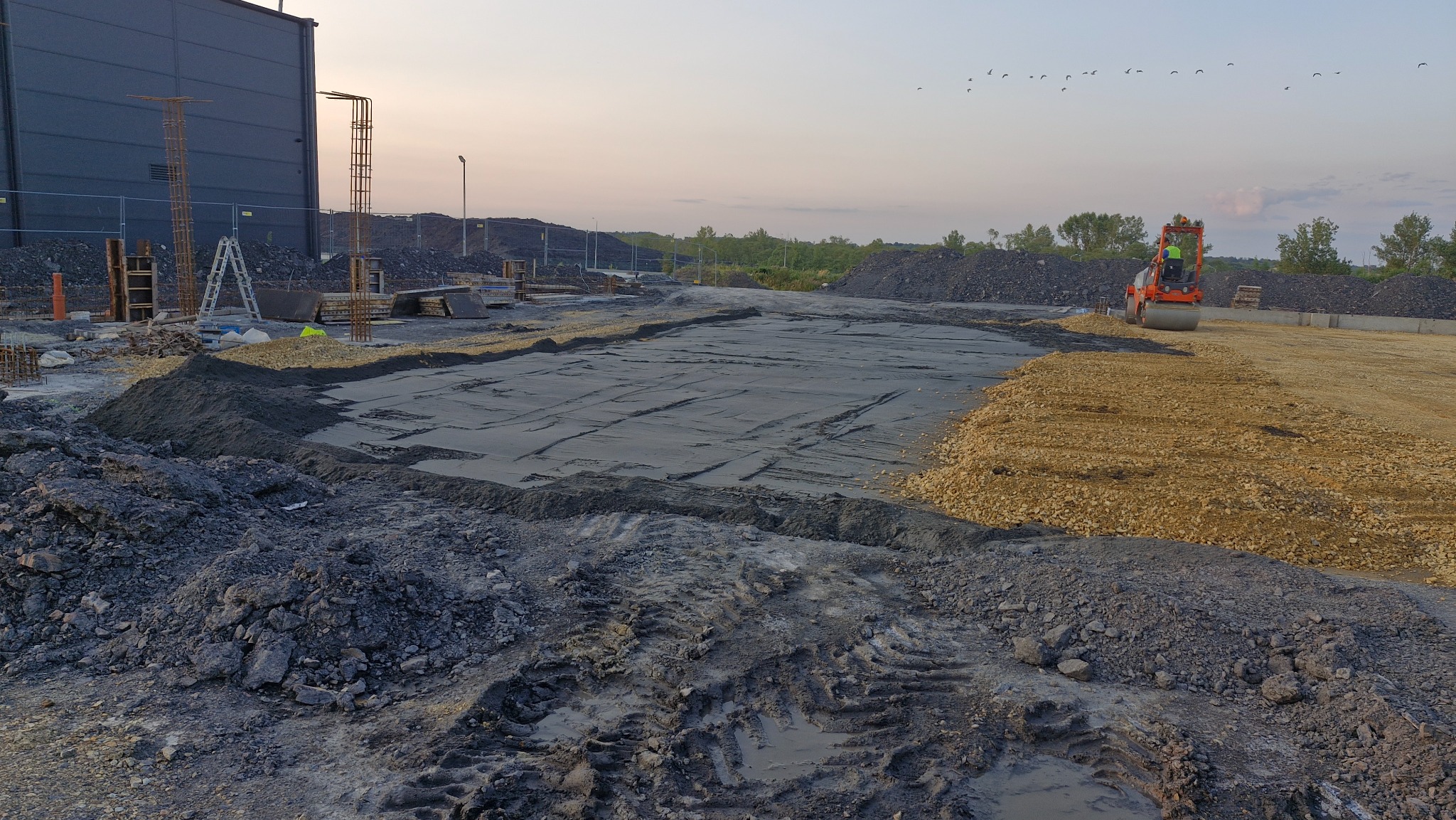 Walec drogowy ubija teren przygotowany pod budowę, widoczne ślady opon w błocie i świeżo ułożony tłuczeń, w tle konstrukcja budynku i sterta ziemi.