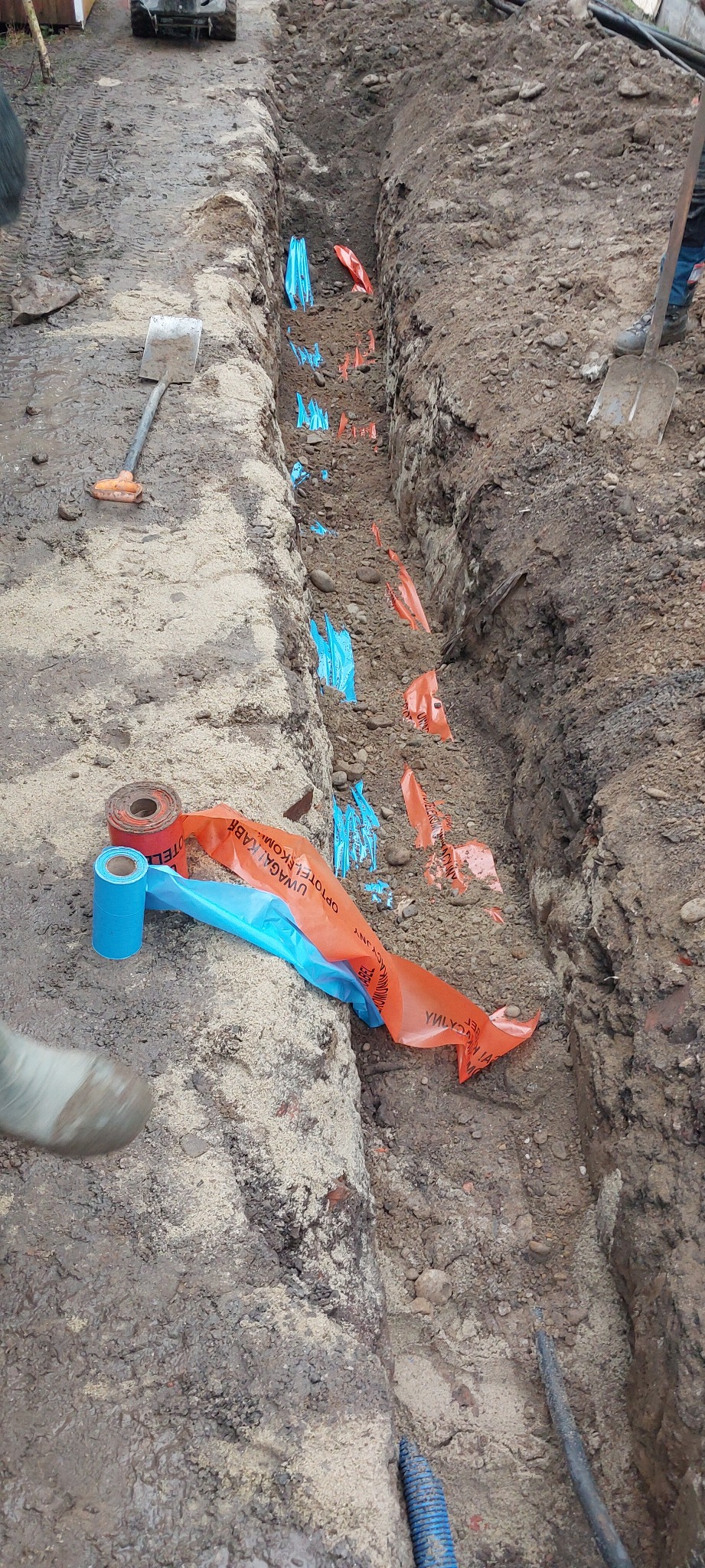 Wykopany wąski rów w ziemi z ułożoną folią ostrzegawczą w kolorze pomarańczowym i niebieskim, widoczne dwie łopaty i fragmenty niebieskiej rury karbowanej.