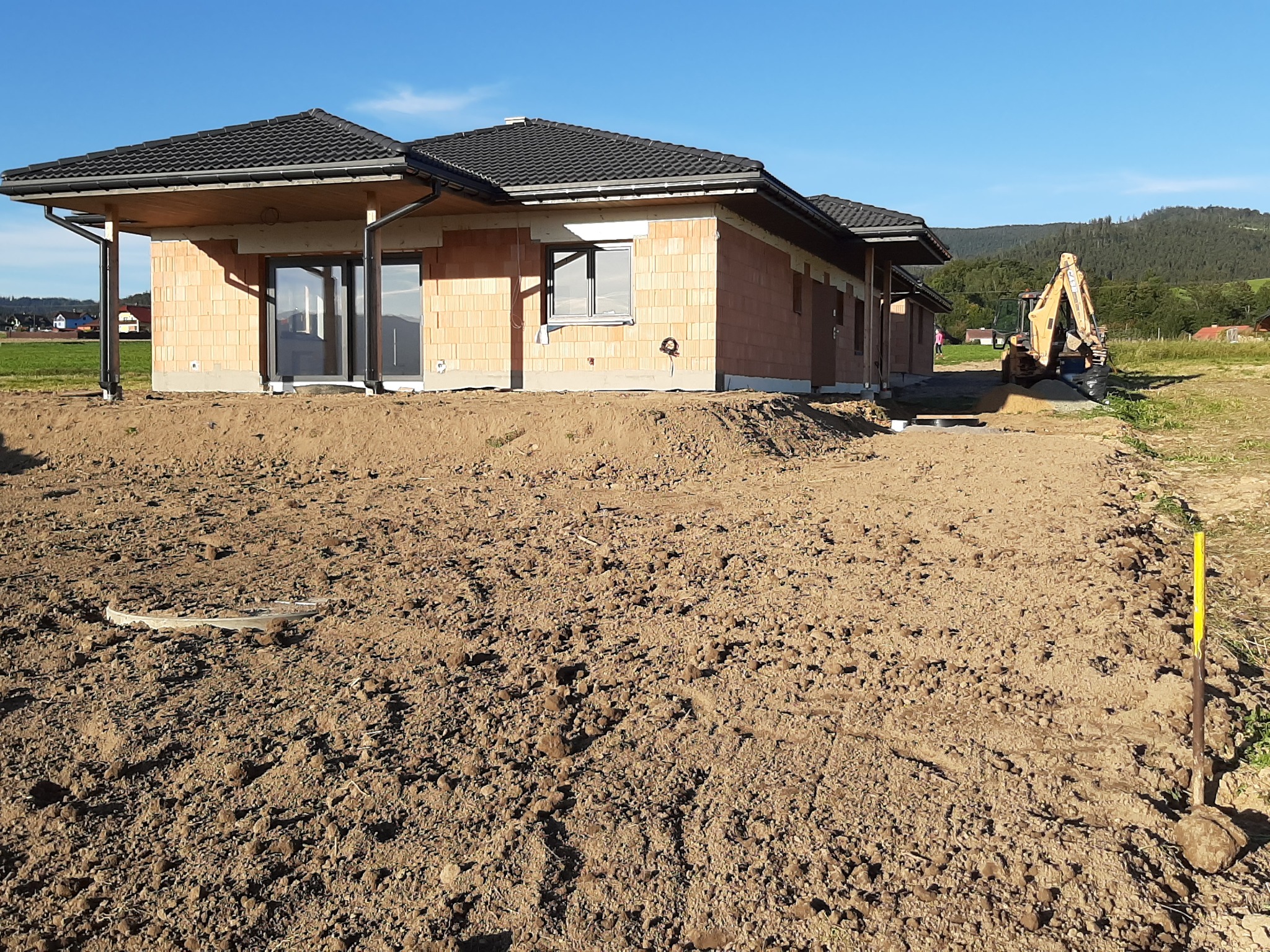 Budynek w stanie surowym otwartym z widoczną konstrukcją dachu i pustakami ceramicznymi, z żółtą koparką w tle na nierównym terenie.