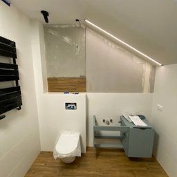 Firma Usługowo-Budowlana - Łazienka w trakcie prac wykończeniowych: częściowo położona drewnopodobna glazura na ścianie, podwieszana toaleta zabezpieczona folią, zamontowana szafka pod umywalkę w kolorze gołębim, widoczne...