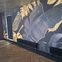 Firma Usługowo-Budowlana - Pokój z ciemną ścianą ozdobioną motywem tropikalnych liści w odcieniach złota i szarości, parkiet w jodełkę, czarne grzejniki i roleta w oknie.