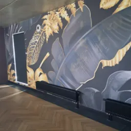 Pok&oacute;j z ciemną ścianą ozdobioną motywem tropikalnych liści w odcieniach złota i szarości, parkiet w jodełkę, czarne grzejniki i roleta w oknie.