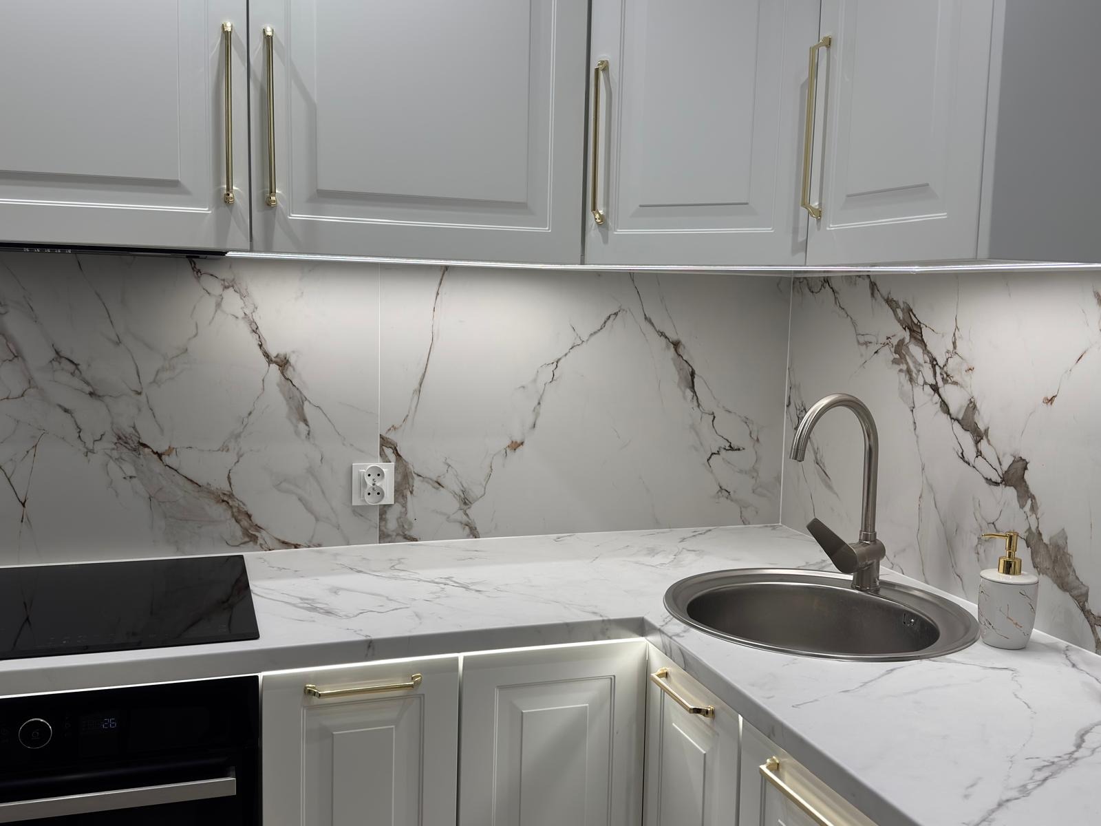 Szara kuchnia z marmurowym blatem i backsplashiem, złote uchwyty, oświetlenie LED pod szafkami, zlewozmywak ze stali nierdzewnej i dozownik mydła.