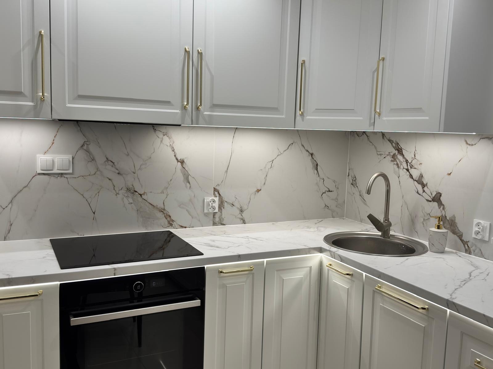Nowoczesna kuchnia z białymi szafkami z frezowanymi frontami, złotymi uchwytami, blatem i ścianą z marmurowym wzorem, oświetleniem LED pod szafkami górnymi i pod blatem, oraz czarną płytą...