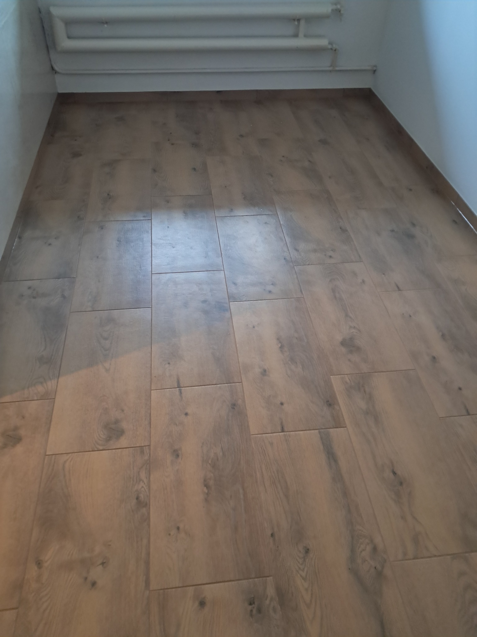 Ułożona podłoga z paneli imitujących drewno w jasnym pomieszczeniu z rurami grzewczymi przy suficie.