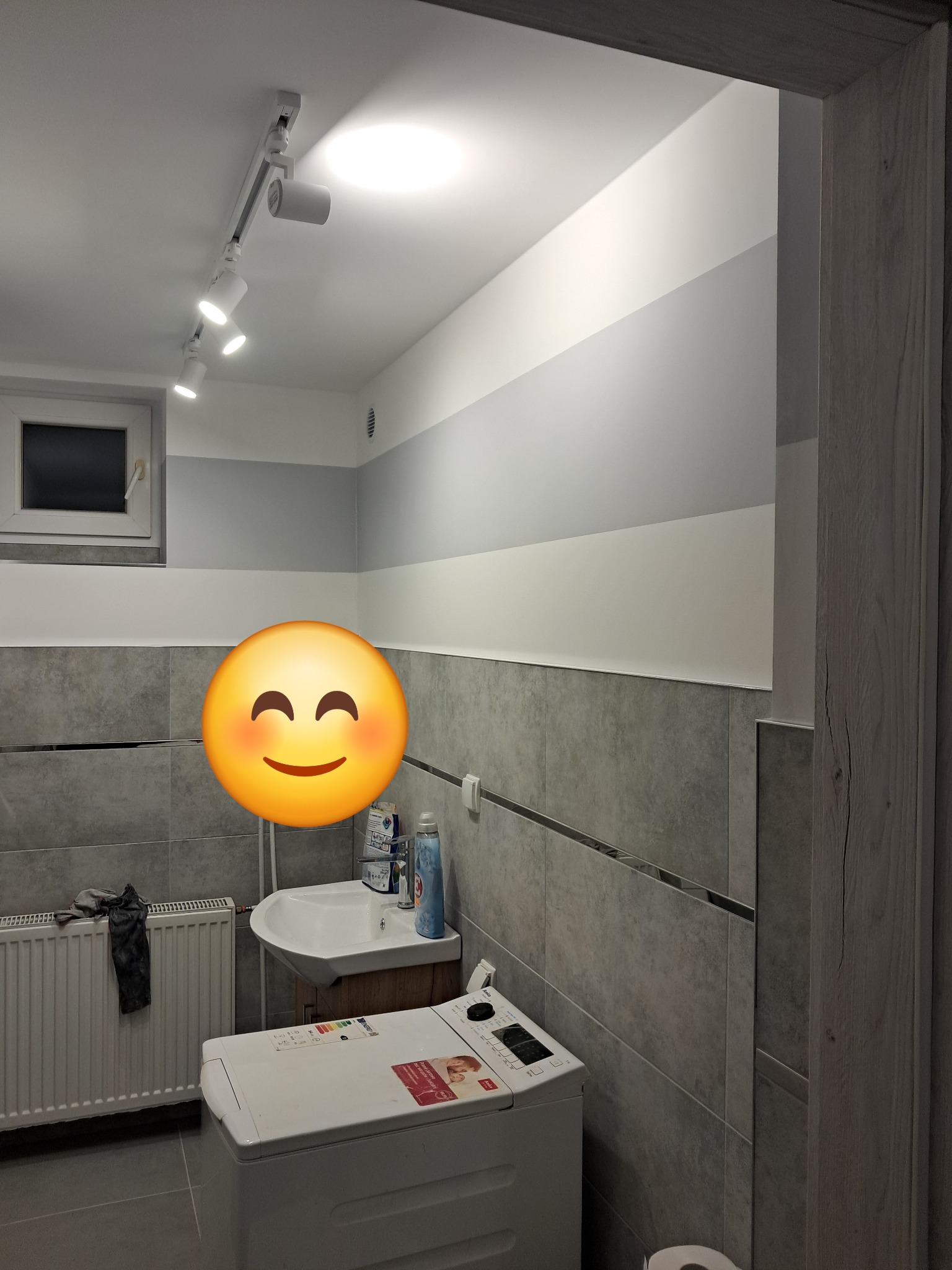Szara łazienka z poziomymi pasami na ścianie, oświetleniem punktowym na suficie, umywalką, grzejnikiem i pralką.