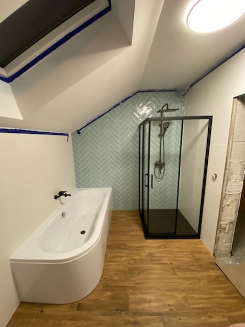 Wykończona łazienka na poddaszu ze skośnym sufitem, z białą wanną narożną, kabiną prysznicową z czarną ramą i błękitnymi płytkami ułożonymi w jodełkę na ścianie.