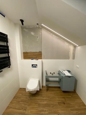 Łazienka w trakcie prac wykończeniowych: częściowo położona drewnopodobna glazura na ścianie, podwieszana toaleta zabezpieczona folią, zamontowana szafka pod umywalkę w kolorze gołębim, widoczne...