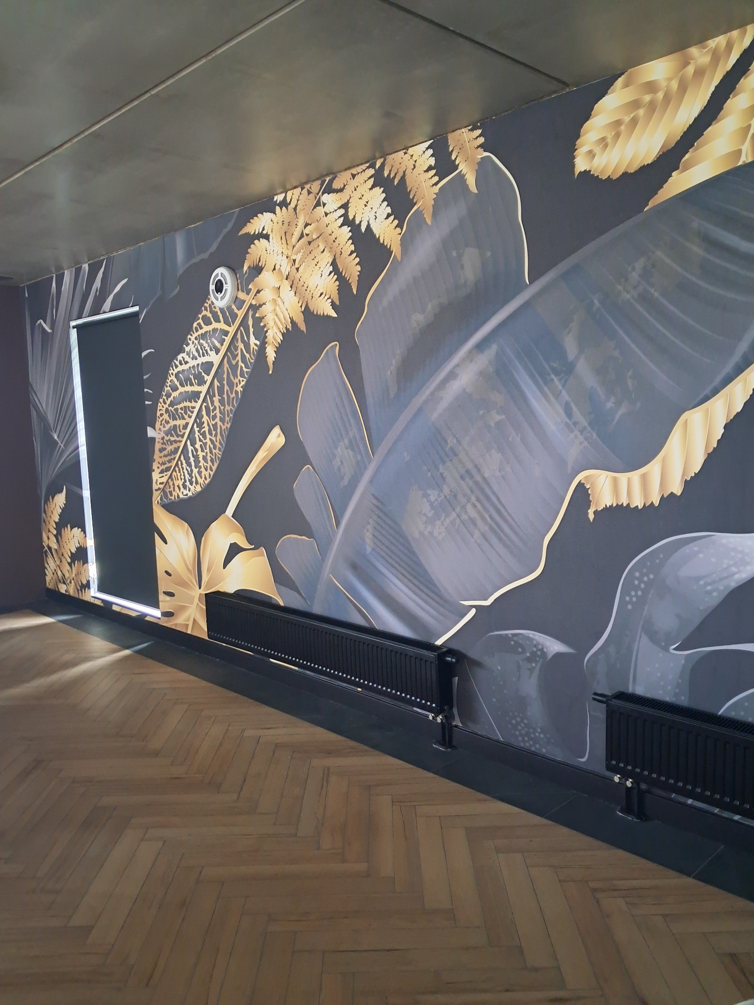 Pokój z ciemną ścianą ozdobioną motywem tropikalnych liści w odcieniach złota i szarości, parkiet w jodełkę, czarne grzejniki i roleta w oknie.