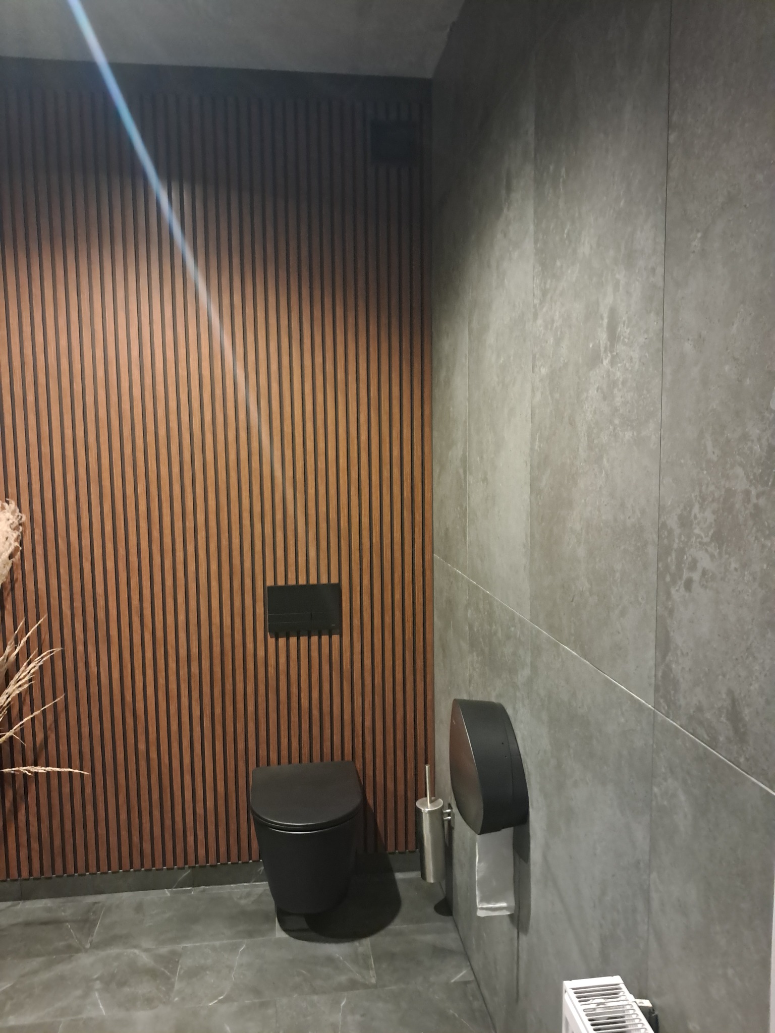 Nowoczesna łazienka z czarną wiszącą toaletą na tle ściany z drewnianych paneli, szare płytki na ścianach i podłodze, minimalistyczny design.