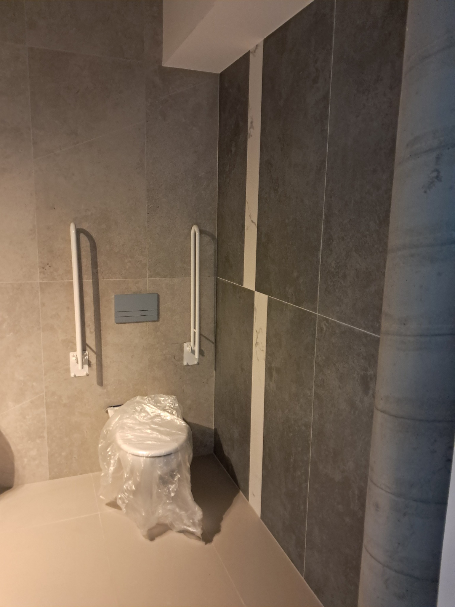 Nowoczesna łazienka z szarymi płytkami, białymi poręczami dla niepełnosprawnych i toaletą zabezpieczoną folią ochronną.