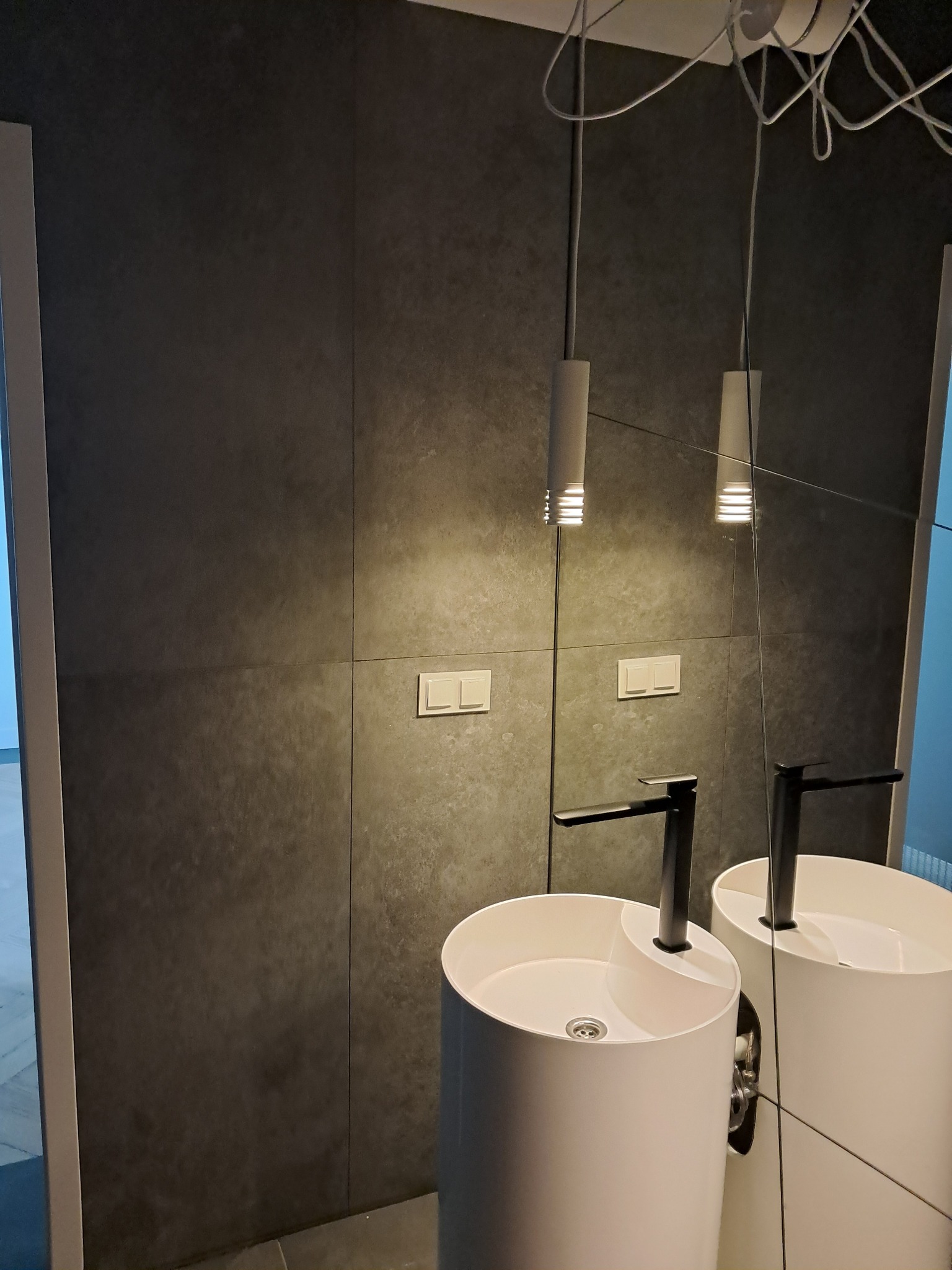 Nowoczesna łazienka z ciemnoszarą okładziną ścienną, białą umywalką wolnostojącą z czarną armaturą i podłużnymi lampami wiszącymi, odbijającymi się w lustrze