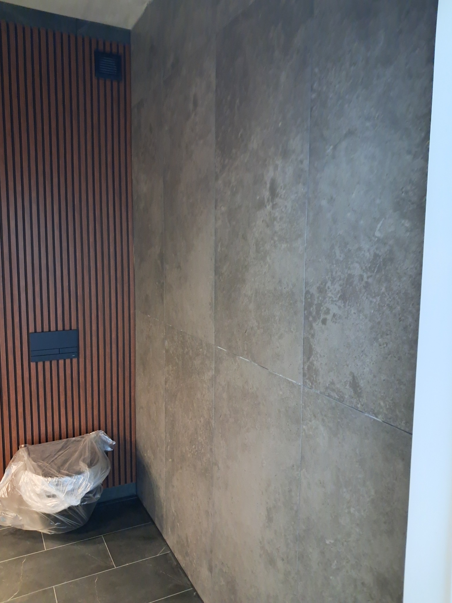 Ściana wyłożona dużymi, szarymi płytkami imitującymi kamień, obok drewniane panele ścienne z ukrytym systemem spłukiwania toalety, na podłodze ciemne płytki, a obok ściany, pod panelami, toaleta...