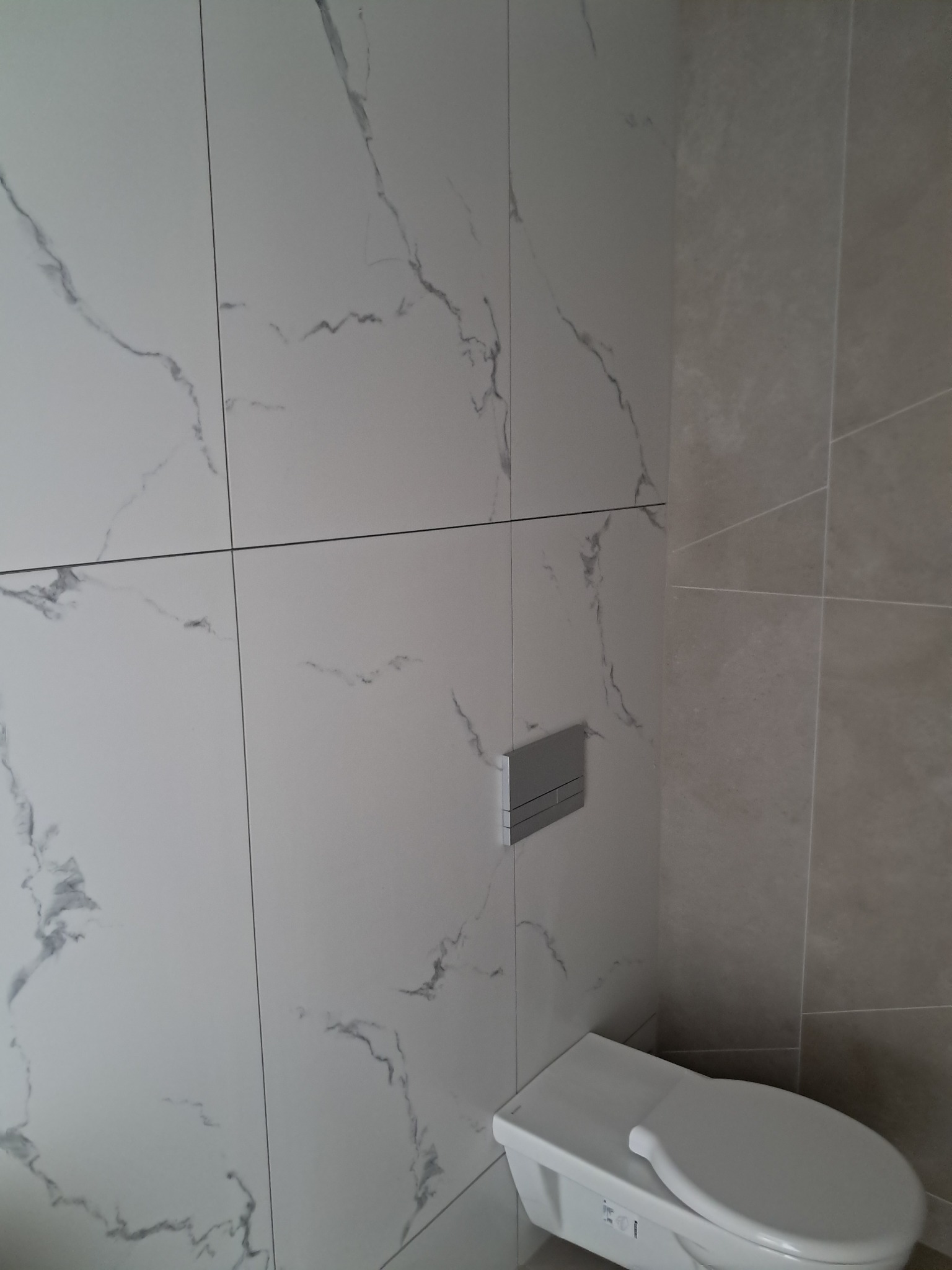 Nowoczesna toaleta z białymi płytkami imitującymi marmur na ścianie i chromowanym przyciskiem spłuczki, widok z boku.