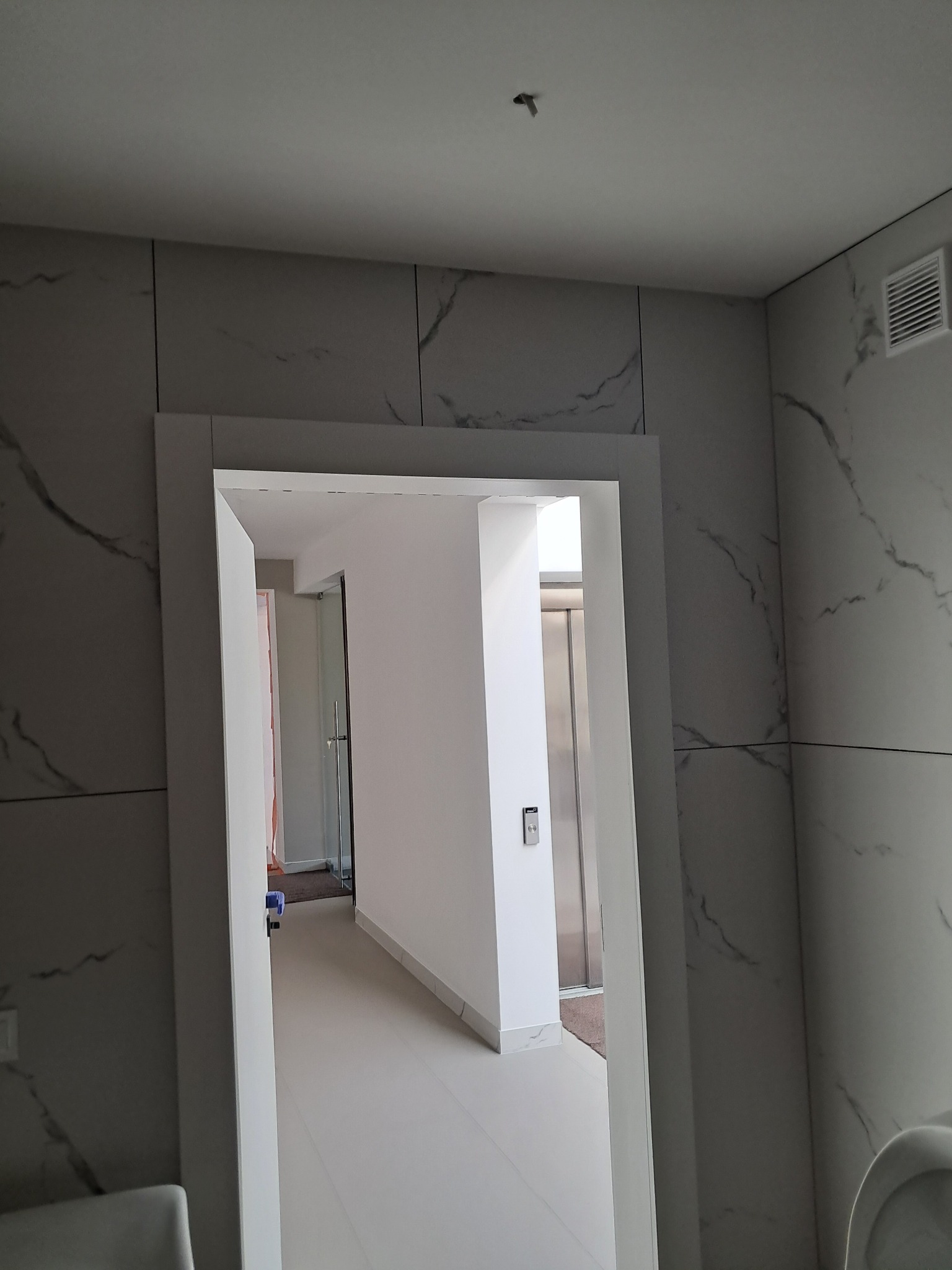 Szare płytki imitujące marmur na ścianie, białe płytki na podłodze, widok przez futrynę drzwi do kolejnego pomieszczenia z windą.