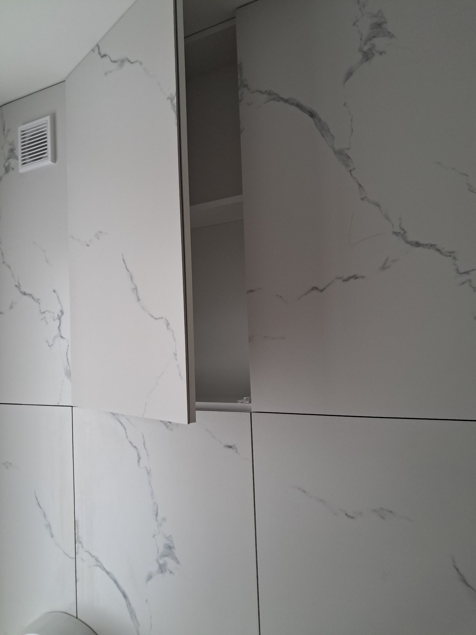 Wykończona ściana w łazience z białych płytek imitujących marmur, z otwartą szafką wiszącą i kratką wentylacyjną.