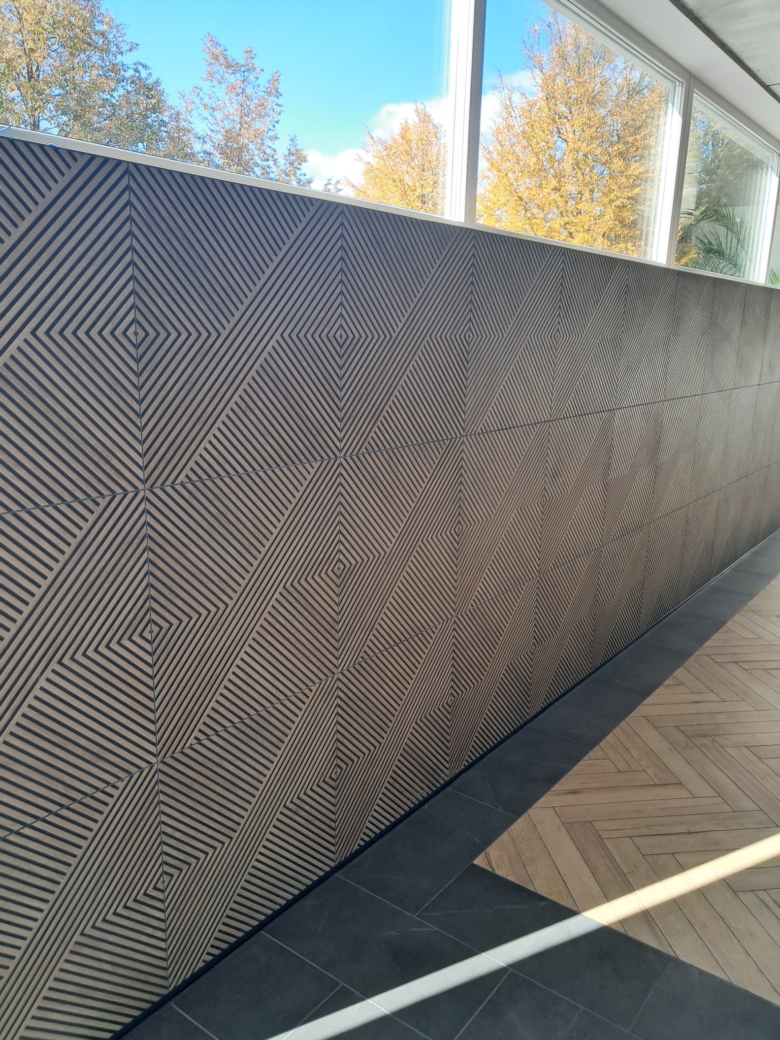 Dekoracyjna ściana z paneli z geometrycznym wzorem rombów, widok z boku, z fragmentem okna i jesiennymi drzewami w tle.