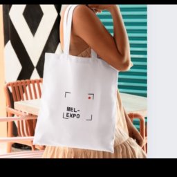 Mel-Expo Emilia Statucka-Sklepik - Biała torba bawełniana z nadrukiem MEL-EXPO, trzymana przez modelkę na tle geometrycznych wzorów i turkusowych paneli.