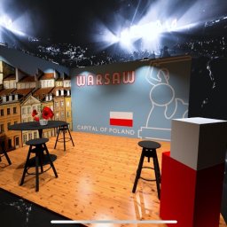 Mel-Expo Emilia Statucka-Sklepik - Scena eventowa z dekoracjami nawiązującymi do Warszawy: tło z wizerunkiem kamienic Starego Miasta, napis 'Warsaw Capital of Poland' oraz stylizowany symbol Warszawskiej Syrenki. Na pierwszym planie...