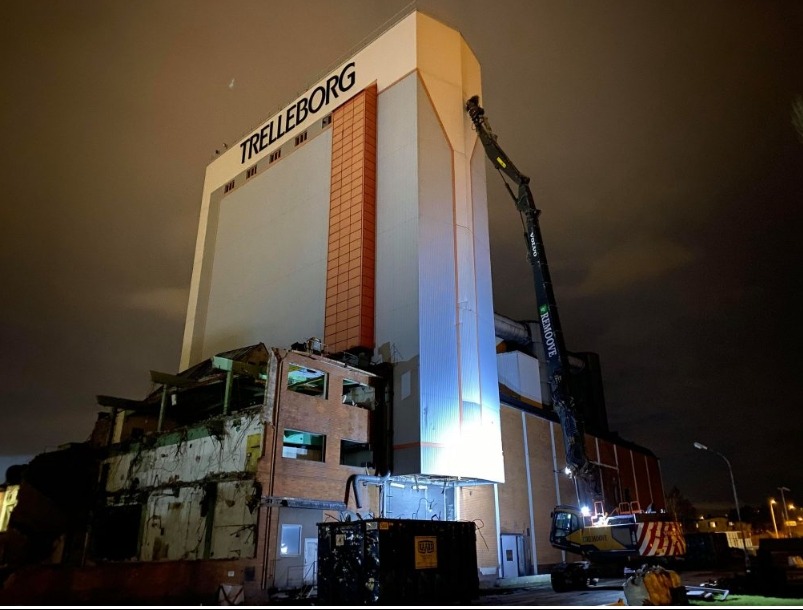 Nocna rozbiórka budynku Trelleborg z użyciem koparki z długim ramieniem, widoczne gruzy i kontener na odpady.