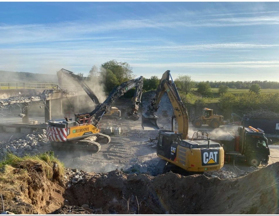 Dwa duże, żółto-czarne koparki hydrauliczne marki Volvo i CAT kruszą betonowy most na tle zielonych pól i błękitnego nieba, z widoczną ciężarówką i mniejszą koparką w tle.