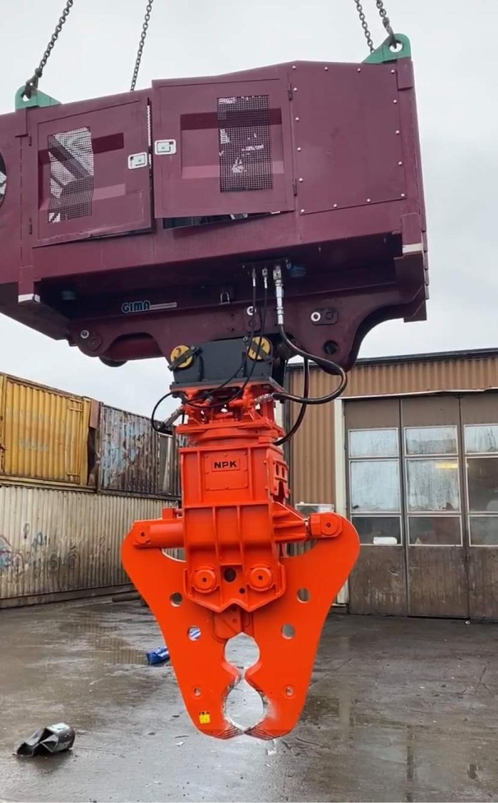 Pomarańczowe nożyce hydrauliczne NPK zawieszone na łańcuchach, gotowe do cięcia metalu, z widocznymi przewodami hydraulicznymi, na tle kontenerów i budynku z oknami.