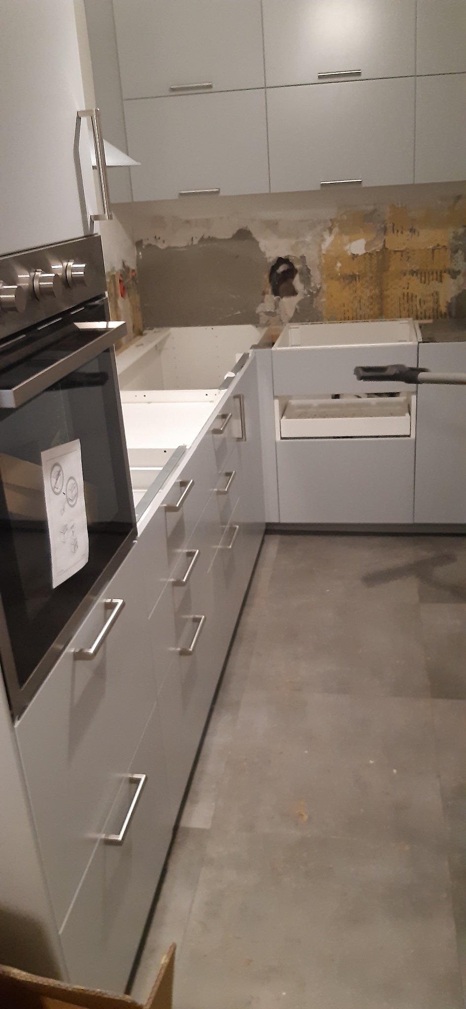 Fragment kuchni w trakcie montażu: szare szafki z podłużnymi uchwytami, wbudowany piekarnik z instrukcją na szybie oraz ściana z widocznymi śladami po poprzednich płytkach.