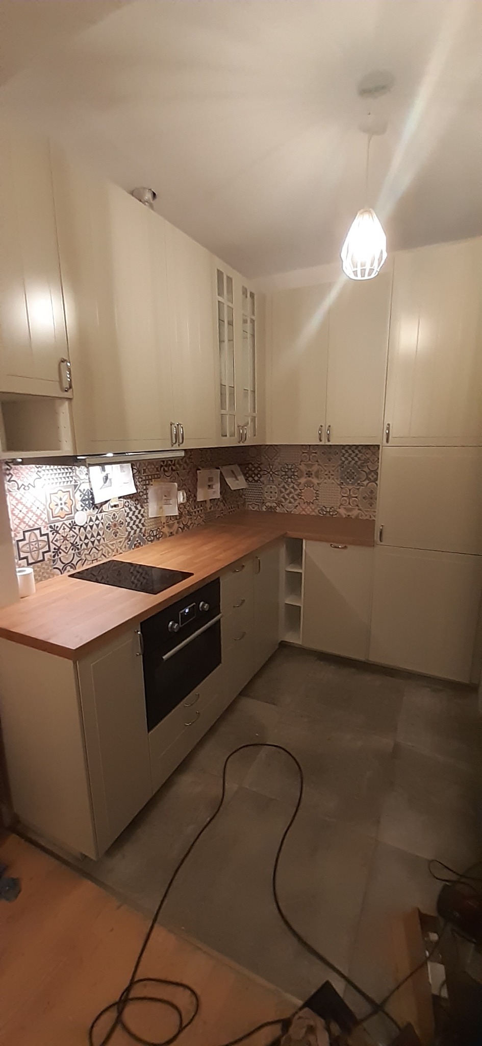 Nowoczesna kuchnia z białymi szafkami, drewnianym blatem i dekoracyjną mozaiką nad blatem, oświetlona wiszącą lampą z ażurowym kloszem.