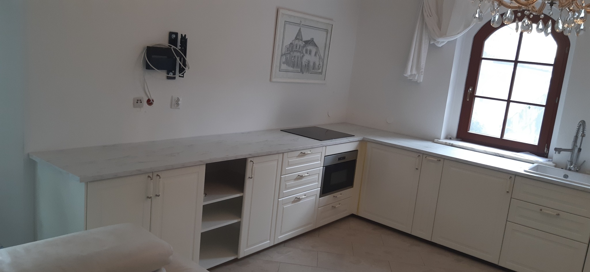 Jasna kuchnia z białymi szafkami i marmurowym blatem pod oknem z brązową ramą, na ścianie wisi obrazek i uchwyt do telewizora.