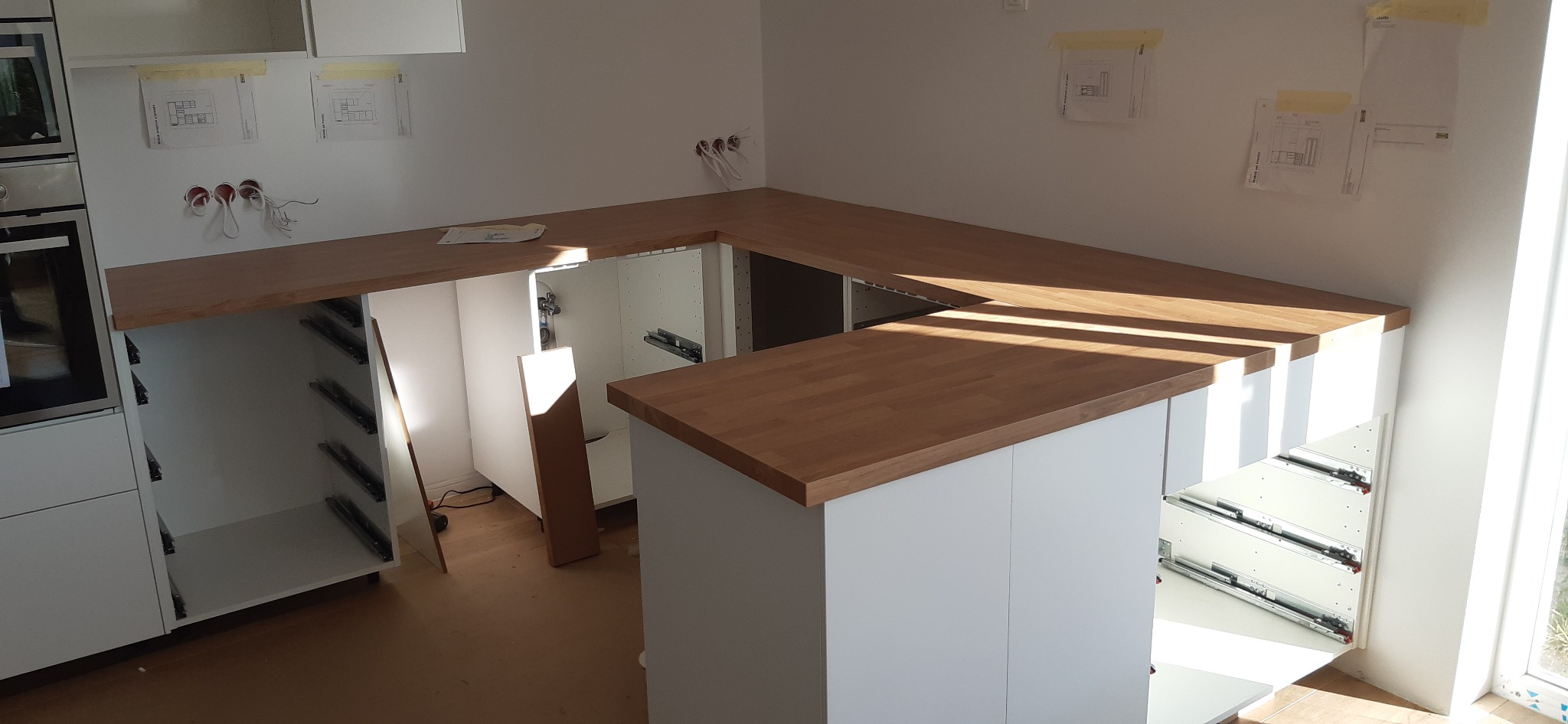 Fragment kuchni w trakcie montażu: białe szafki z drewnianym blatem, widoczne szuflady na prowadnicach, plany na ścianie, instalacja elektryczna.