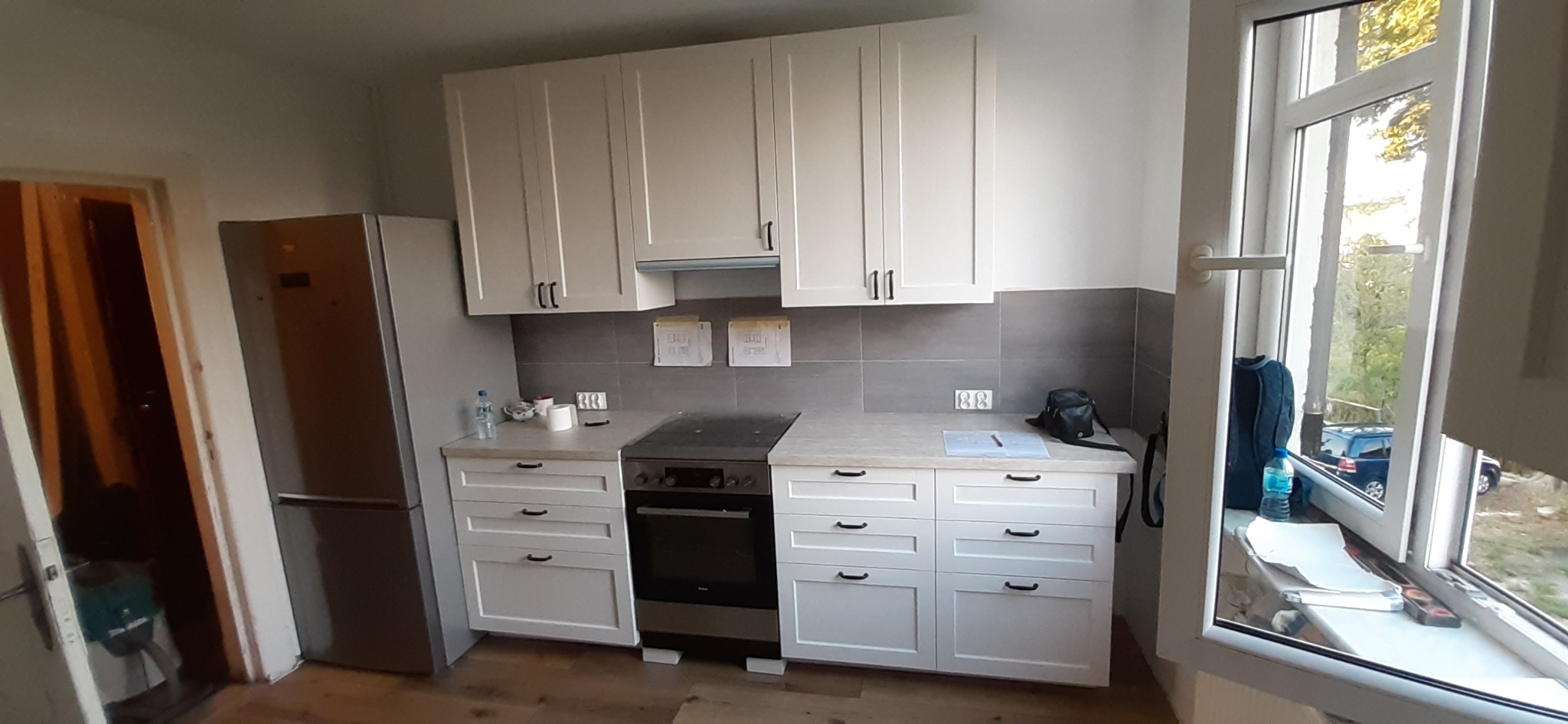 Biała zabudowa kuchenna z górnymi szafkami i szufladami dolnymi, szarym pasem płytek nad blatem, widoczne plany na ścianie, obok okna plecak i butelka wody.