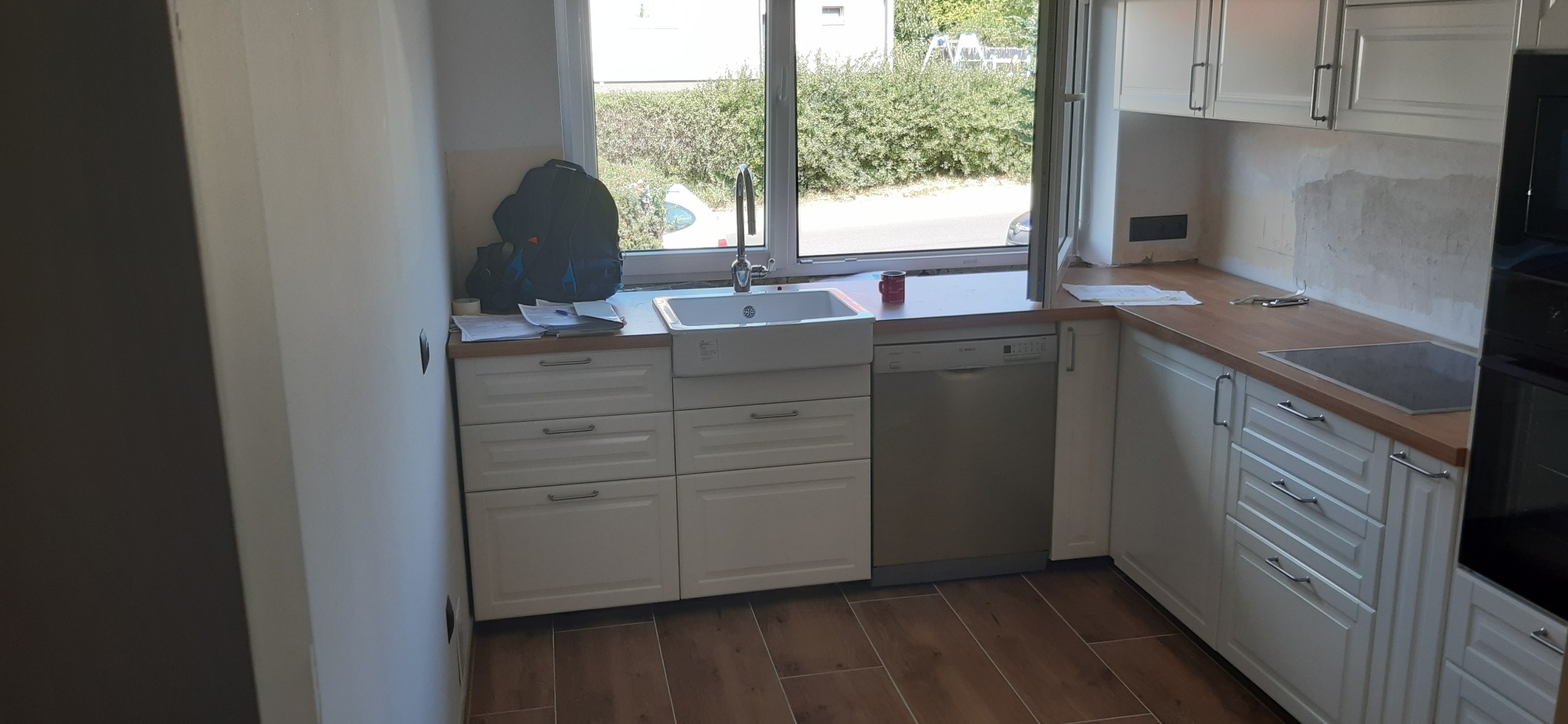 Częściowo zamontowana biała kuchnia w Gryfinie z drewnianym blatem, widoczna zlew, zmywarka i płyta grzewcza. Na blacie leżą narzędzia, a pod oknem stoi plecak.