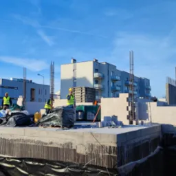 Budowa domu z betonu komórkowego, widoczni pracownicy w żółtych kamizelkach i kaskach, żuraw podnosi elementy szalunku, w tle budynki mieszkalne.