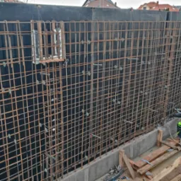Pracownik w odblaskowej kamizelce i niebieskim kasku montuje zbrojenie ściany fundamentowej z czarną szalunką, widoczne elementy mocujące i konstrukcja zbrojeniowa.