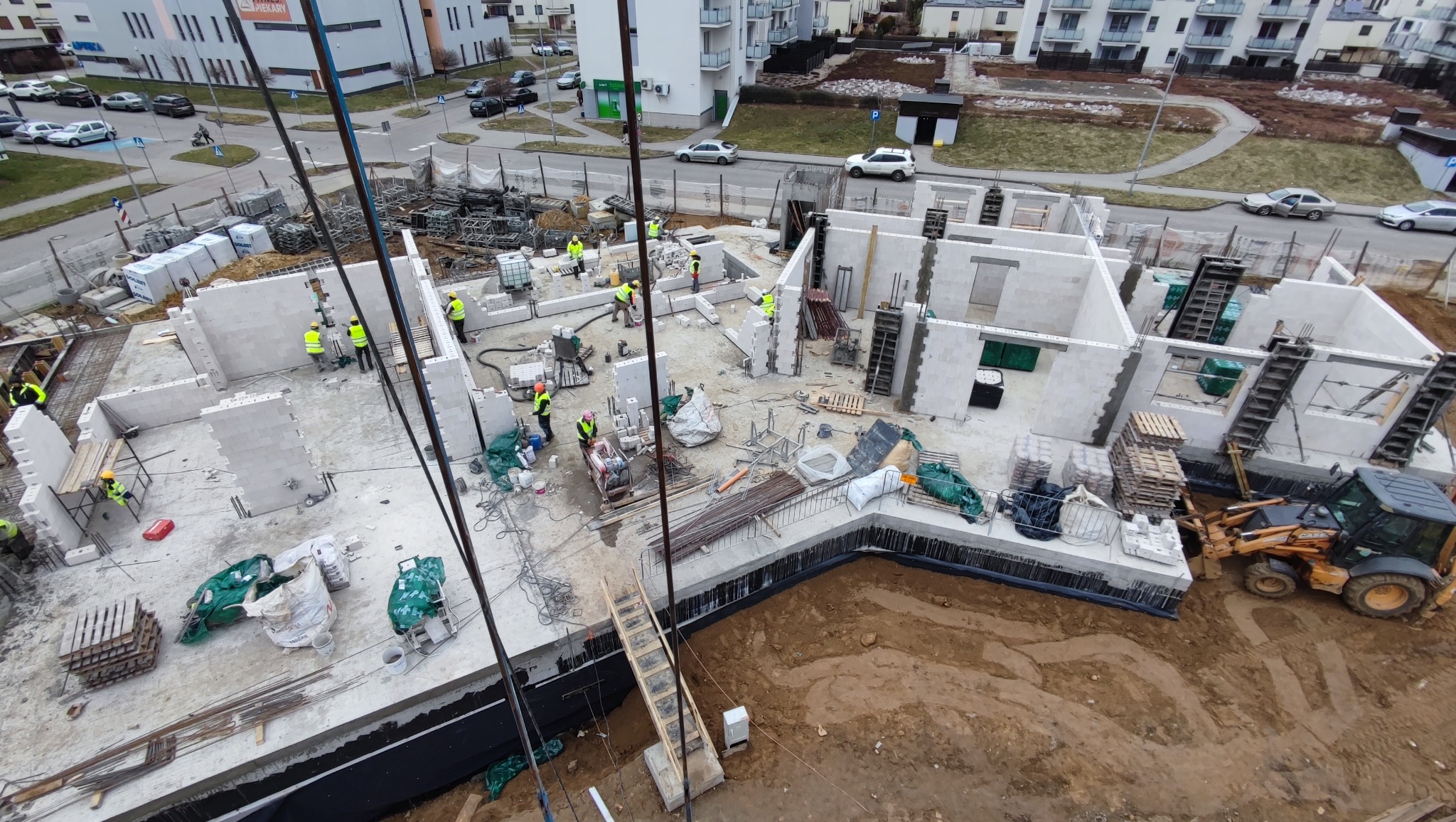 Budowa wielorodzinnego budynku w stanie surowym otwartym z garażem podziemnym: widok z góry na fundamenty, ściany z betonu komórkowego, pracowników w żółtych kamizelkach, palety z materiałami...