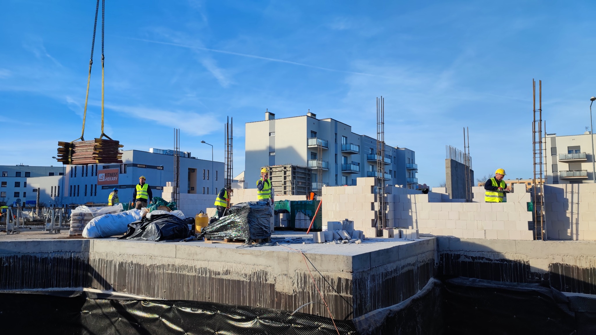 Budowa domu z betonu komórkowego, widoczni pracownicy w żółtych kamizelkach i kaskach, żuraw podnosi elementy szalunku, w tle budynki mieszkalne.