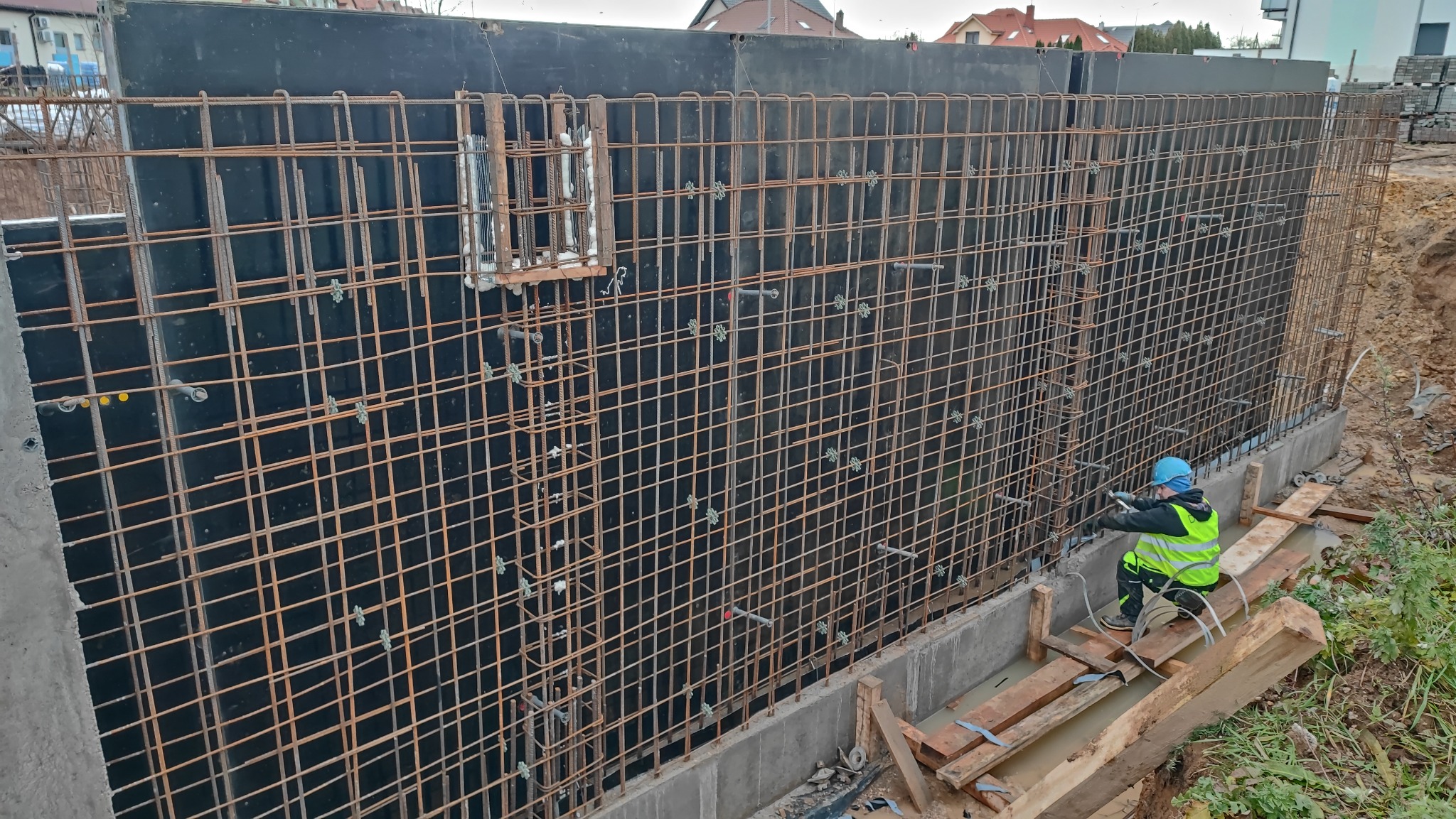 Pracownik w odblaskowej kamizelce i niebieskim kasku montuje zbrojenie ściany fundamentowej z czarną szalunką, widoczne elementy mocujące i konstrukcja zbrojeniowa.