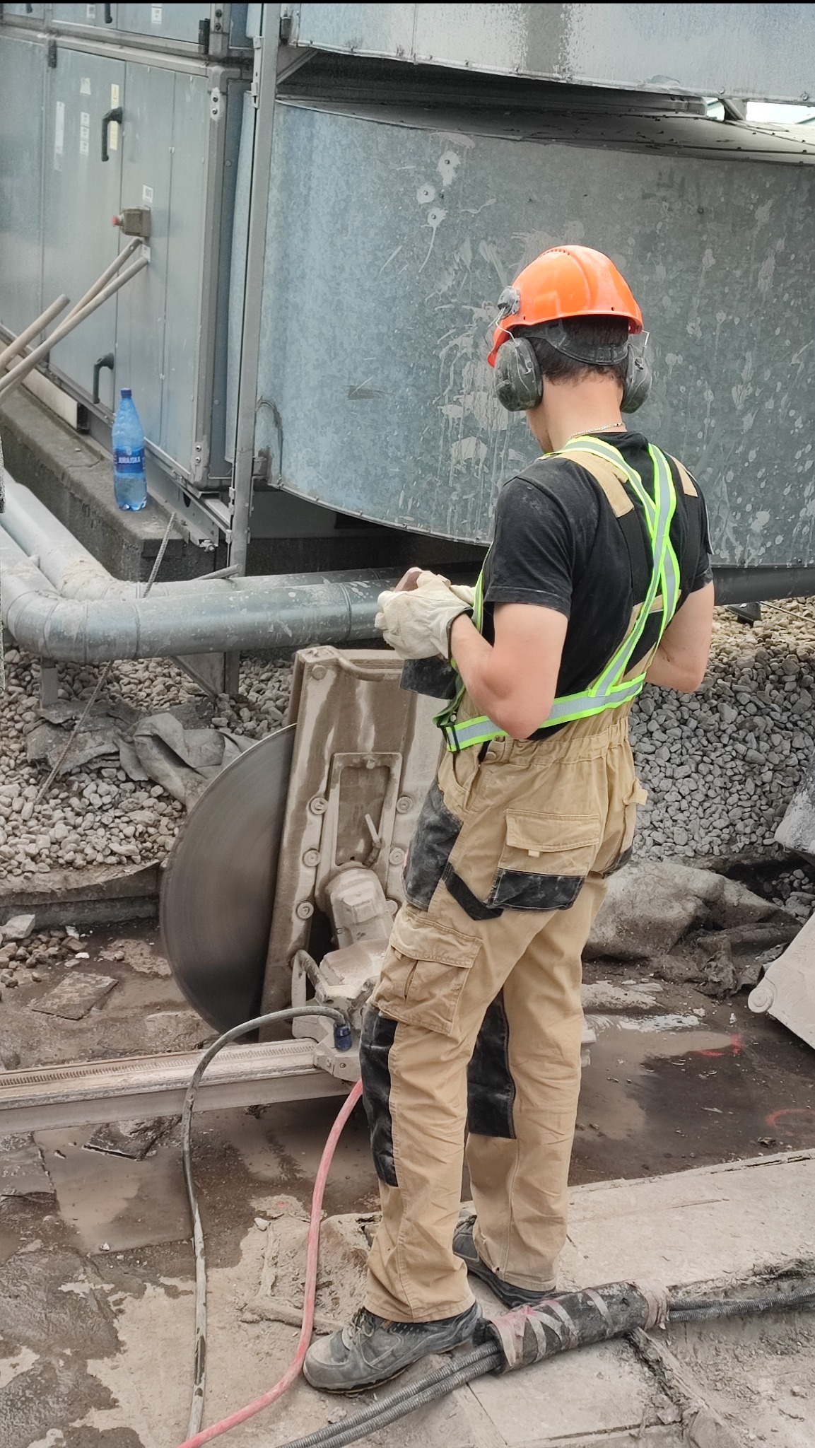 Pracownik w kasku i słuchawkach ochronnych obsługuje piłę diamentową do cięcia betonu na budowie, widoczne elementy wentylacji i gruz.