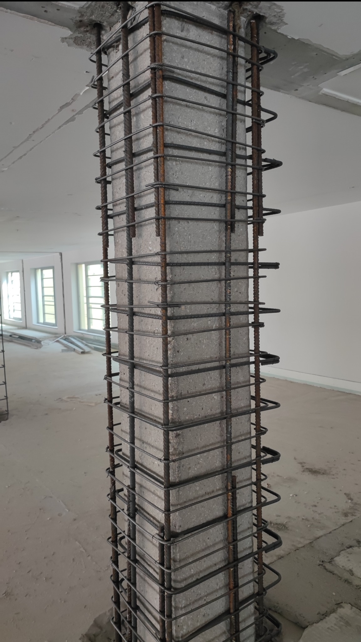 Żelbetowy słup z wystającą stalową siatką zbrojeniową na placu budowy, widoczne okna w tle.