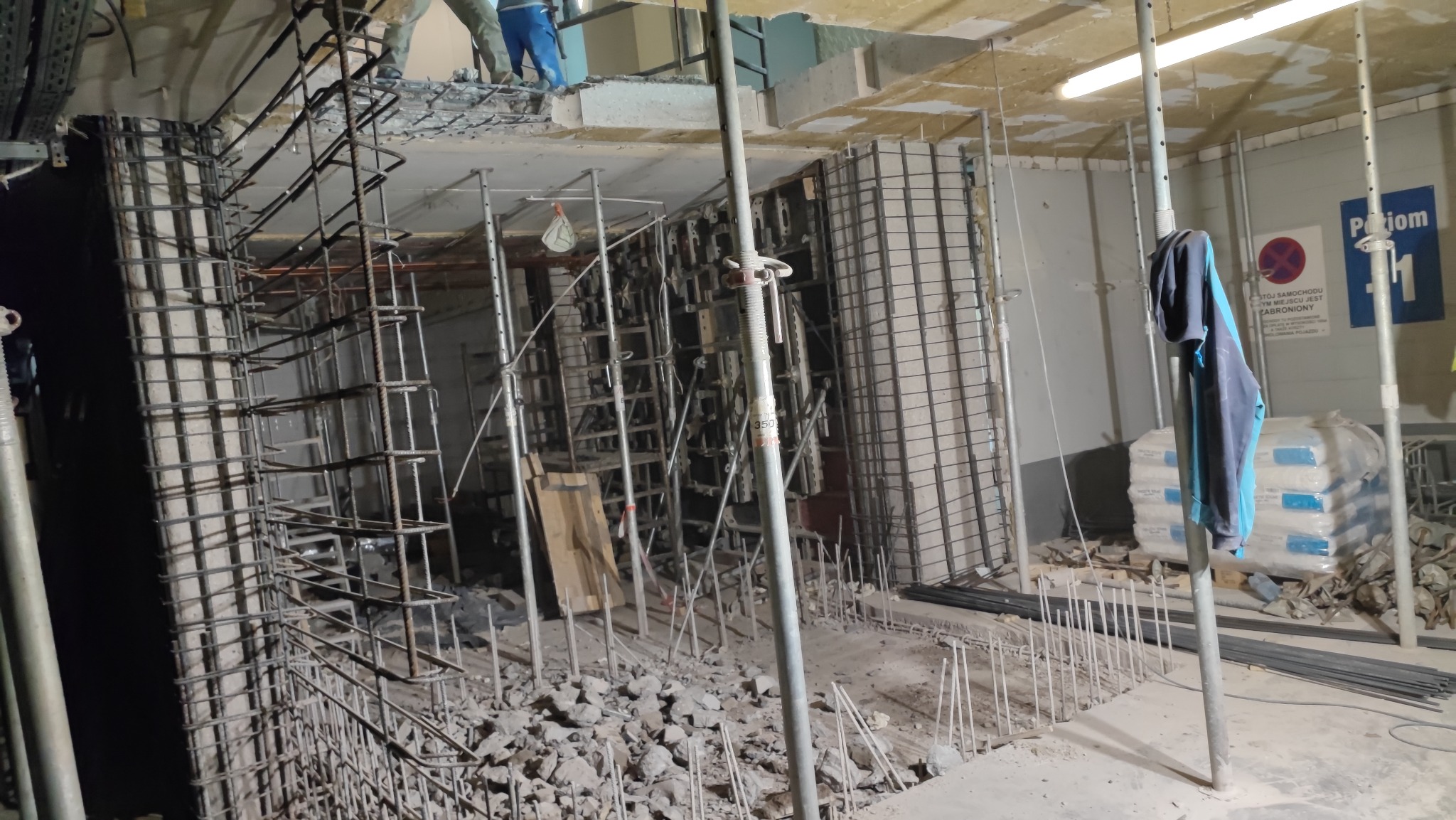 Wnętrze w trakcie budowy z widoczną konstrukcją zbrojeniową, szalunkami, stemplami i stertą gruzu na podłodze; w tle widoczne worki z zaprawą i tablica informacyjna.