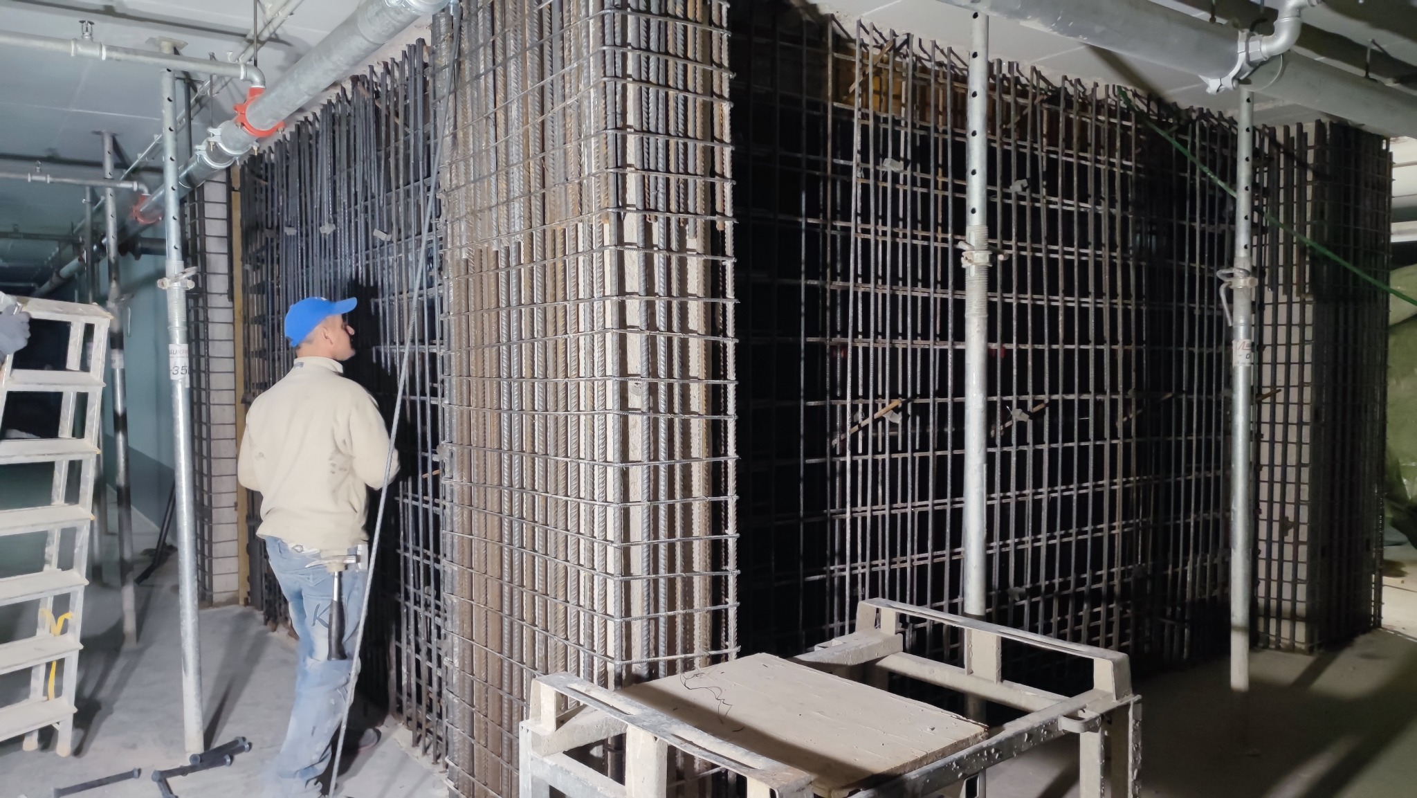Pracownik w niebieskiej czapce przy zbrojeniu słupów i ścian wewnątrz budynku, widoczne szalunki i podpory.