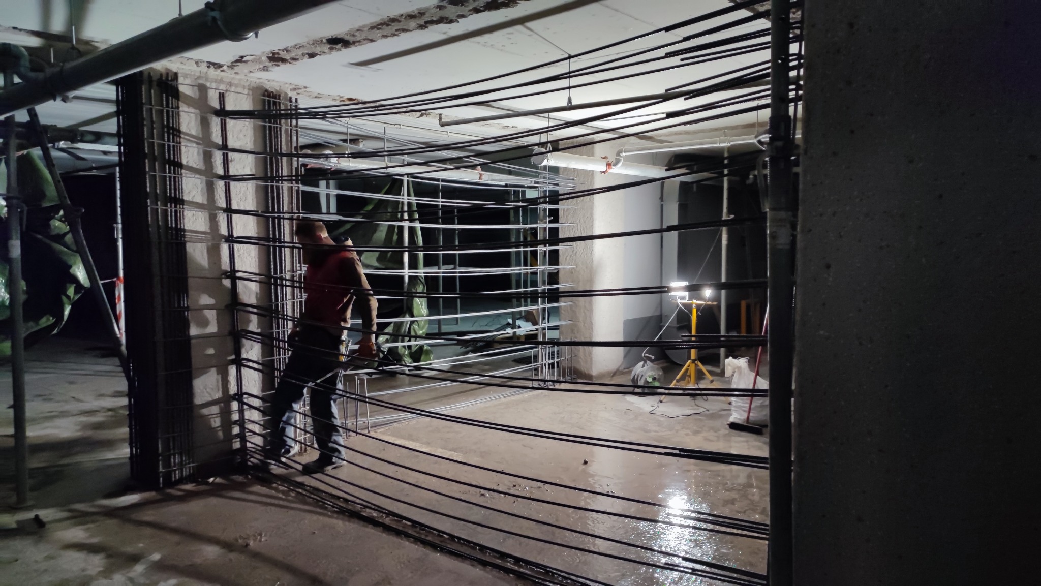 Prace zbrojarskie wewnątrz budynku, widoczna stalowa konstrukcja zbrojeniowa i pracownik w czerwonej koszuli, oświetlenie budowlane w tle.