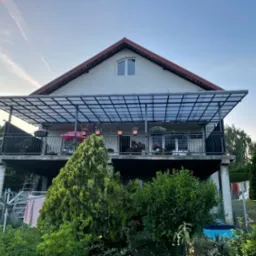 Dwupiętrowy dom z balkonem z metalowymi balustradami i zadaszeniem z przezroczystego tworzywa sztucznego, widok z ogrodu, z elementami prania na suszarce.