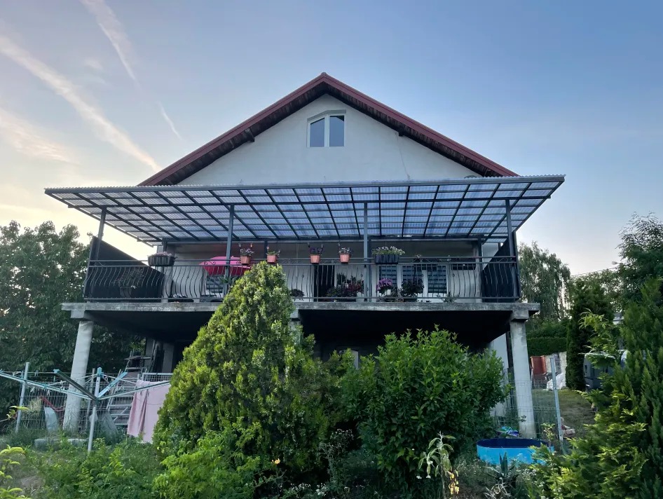 Dwupiętrowy dom z balkonem z metalowymi balustradami i zadaszeniem z przezroczystego tworzywa sztucznego, widok z ogrodu, z elementami prania na suszarce.