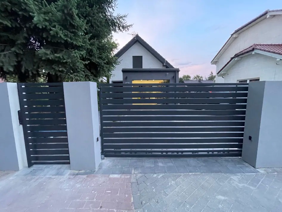 Nowoczesna, pozioma brama wjazdowa i furtka z metalowymi przęsłami w kolorze antracytowym, osadzone na szarych, murowanych słupkach, na tle domu i zielonego drzewa.