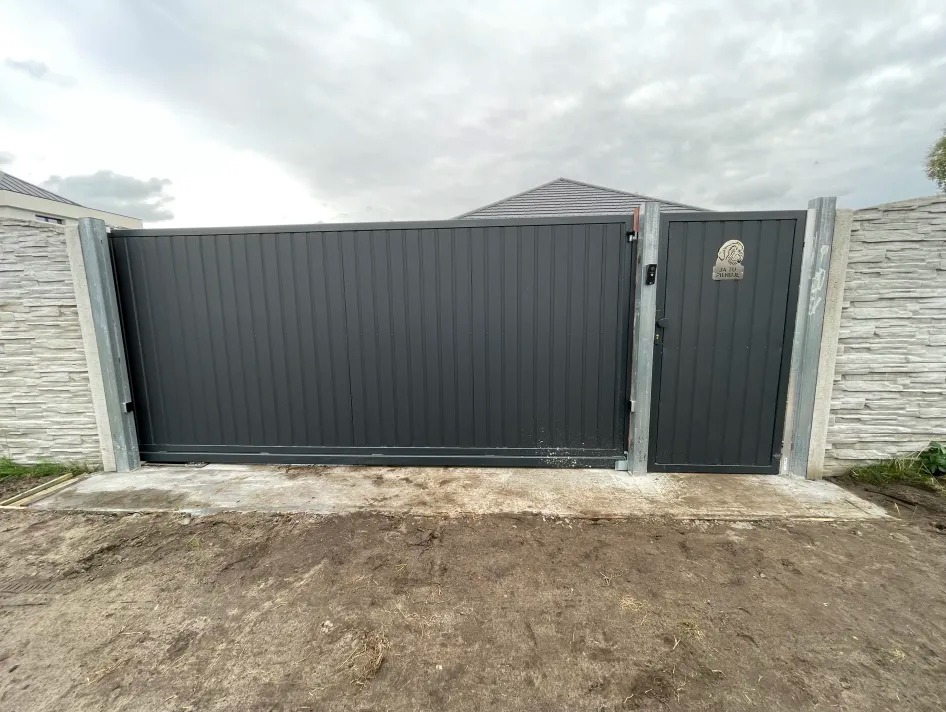 Przesuwna brama metalowa w kolorze grafitowym, z furtką i tabliczką z wizerunkiem psa, zamontowana między betonowymi słupkami z dekoracyjną fakturą kamienia.