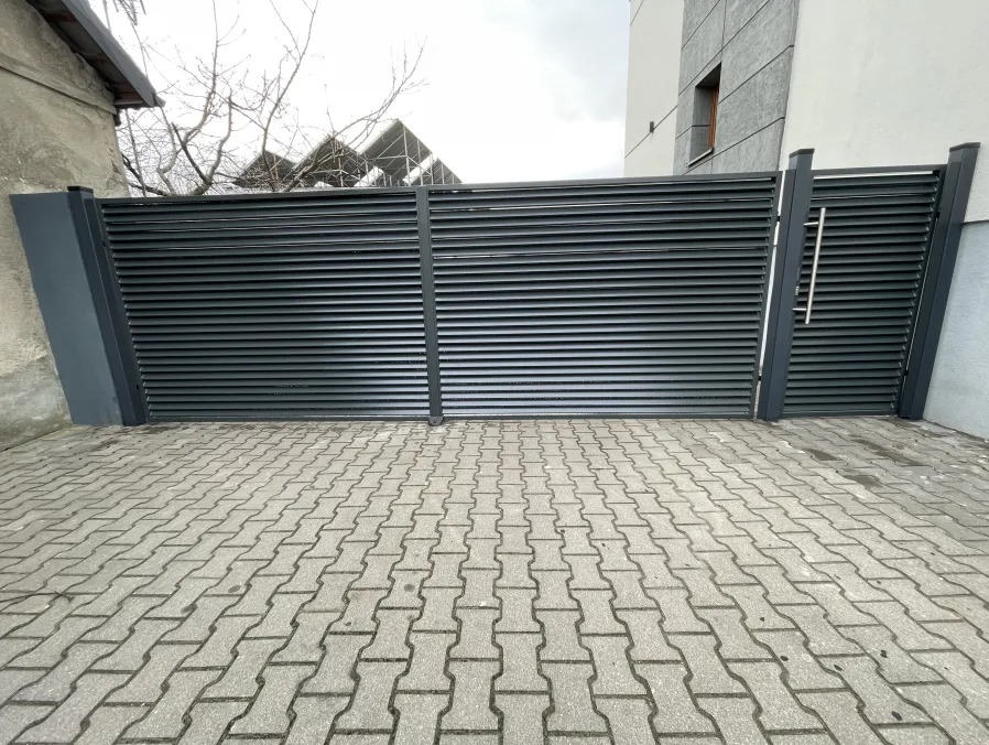 Nowoczesna, pozioma brama przesuwna z metalowych profili w kolorze grafitowym, z widoczną klamką ze stali nierdzewnej, na tle kostki brukowej i fragmentu budynku.