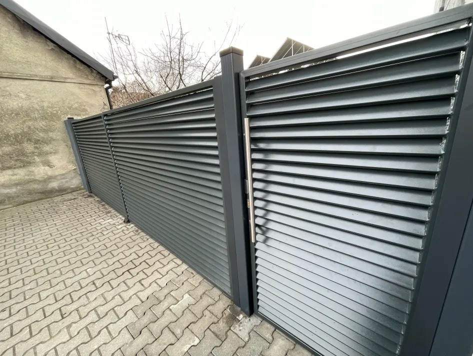 Nowoczesne, szare ogrodzenie lamelowe z bramą, widoczne z boku na tle kostki brukowej i budynku.