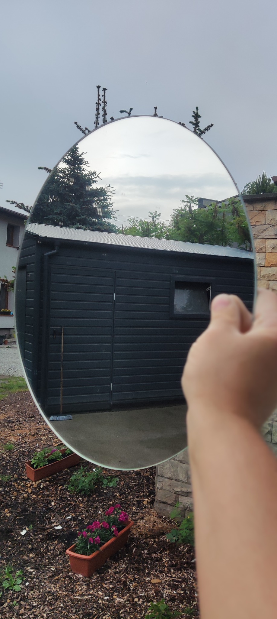 Okrągłe lustro odbija obraz szarego budynku gospodarczego z czarnymi deskami elewacyjnymi, widoczne kwiaty w donicach i fragment kamiennego muru.