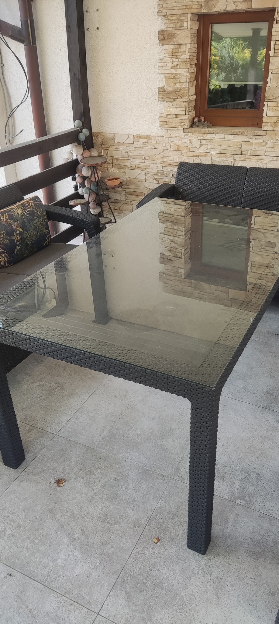 Szklany stół na tarasie z rattanowym wykończeniem, widoczne krzesła i dekoracyjna ściana z kamienia.